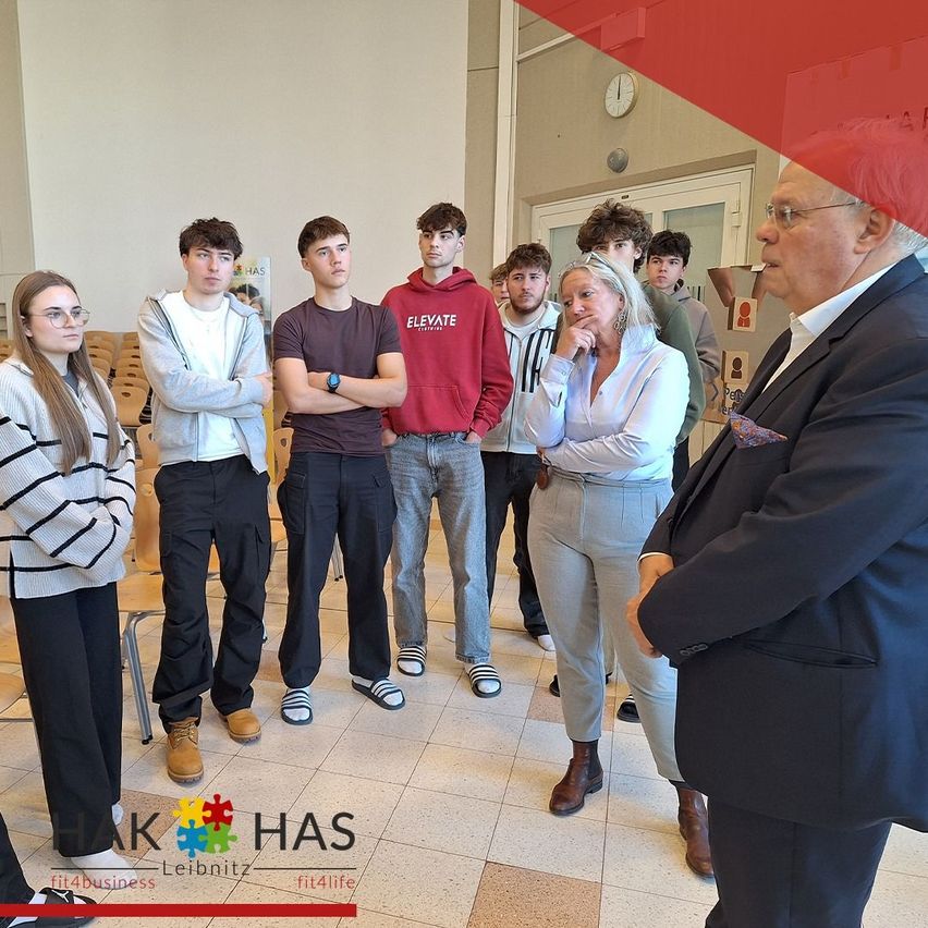 Schüler und Erwachsene versammeln sich in einem Flur, wahrscheinlich für eine Schulveranstaltung. Ein Mann in Anzug spricht mit einer Frau in einer weißen Jacke. Hinter ihnen stehen Schüler in einer Reihe. Der Flur hat weiße Wände, eine Uhr und Türen.