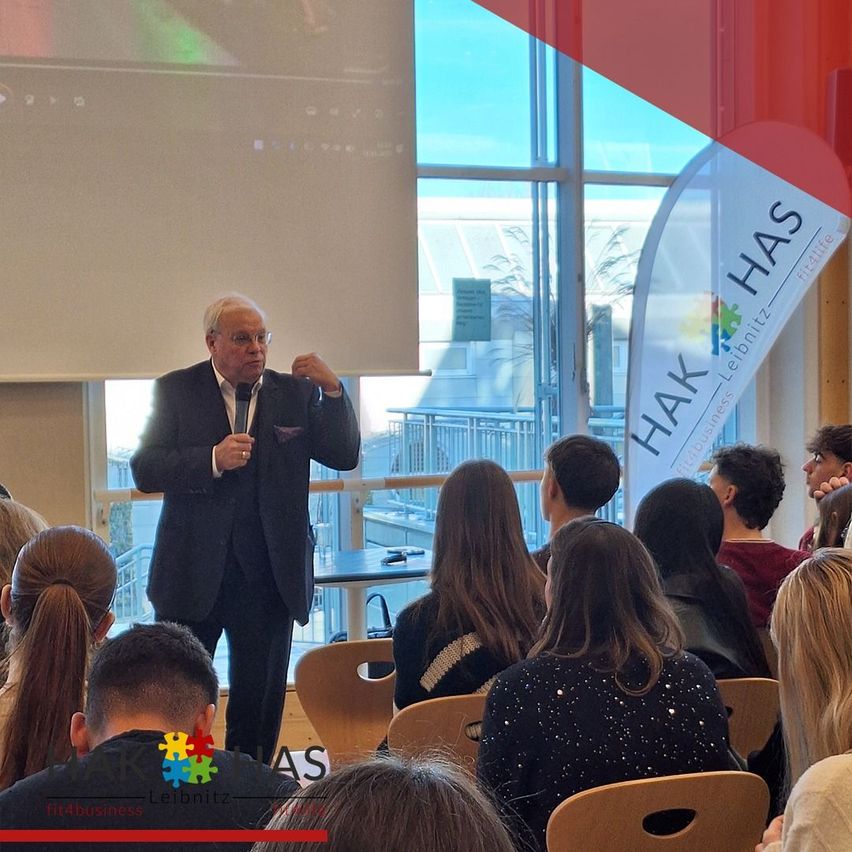 Ein Mann spricht in ein Mikrofon vor einer Projektionsleinwand, mit einem Banner, das 'HAK HAS' liest. Mehrere Menschen sitzen auf Stühlen und hören ihm zu.