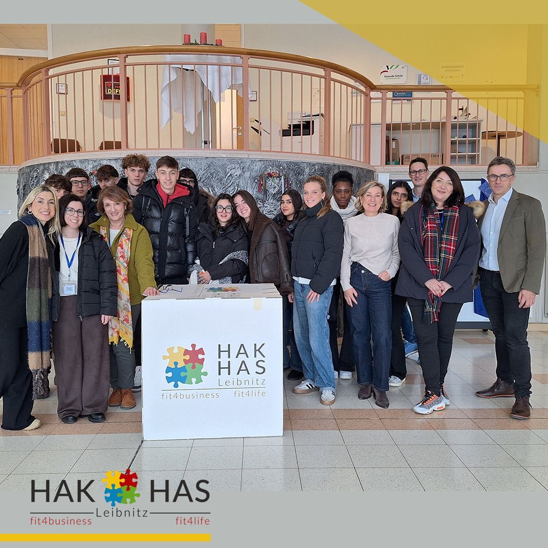 Eine Gruppe von Erwachsenen und Jugendlichen steht lächelnd zusammen in einem modernen Gebäude. Ein weißer Tisch mit einem Logo für HAK HAS Leibniz steht im Vordergrund.