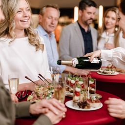 Eine Gruppe von Menschen sitzt um einen Tisch herum, lächelt und plaudert. Eine Person schenkt Champagner in ein Glas. Der Tisch ist mit Tellern mit Essen, Gläsern und einer Flasche Champagner gedeckt.