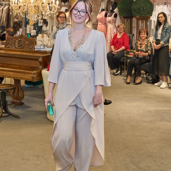 Eine Frau in einem hellgrauen Jumpsuit steht in einem Bekleidungsgeschäft, lächelt und hält eine grüne Handtasche. Hinter ihr sitzen mehrere Frauen auf Stühlen, und links steht ein Klavier.