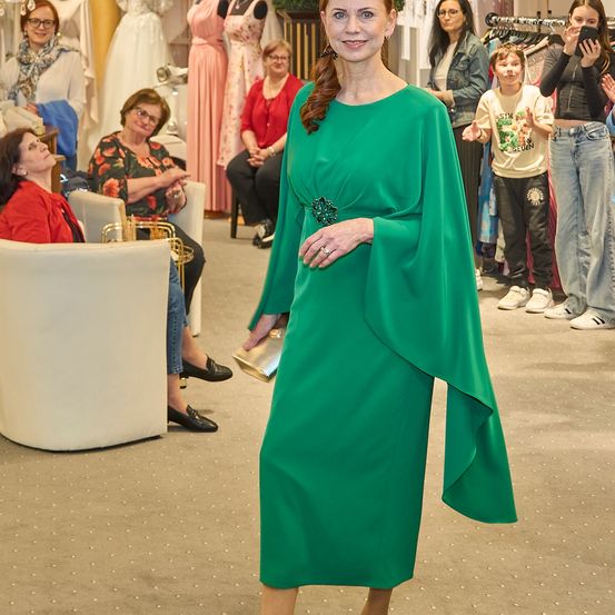 Eine Frau in einem grünen Kleid steht in einem Bekleidungsgeschäft, umgeben von Menschen und Kleiderständern.