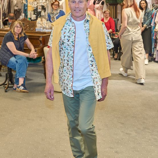 Ein Mann in einem Blumenhemd und gelber Weste geht durch einen Bekleidungsladen, im Hintergrund sind mehrere Personen zu sehen.
