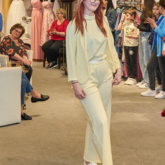 Eine Frau in einem cremefarbenen Outfit geht an einer Gruppe von Menschen in einem Bekleidungsgeschäft vorbei, mit Mannequins und Kleiderständern im Hintergrund.