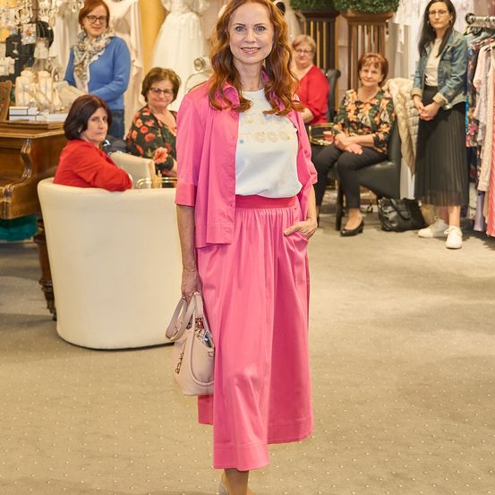 Eine Frau in einem rosa Outfit steht in einem Bekleidungsgeschäft, lächelt und hält eine Handtasche. Hinter ihr sitzen und stehen mehrere Frauen, einige mit Brille.