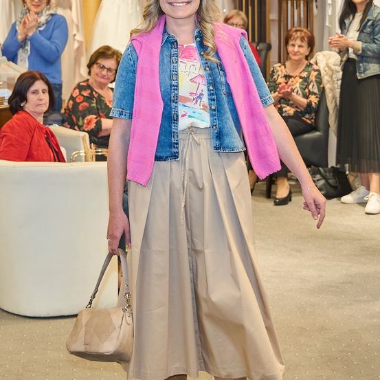 Eine Frau in einem rosa Weste über einer Jeansjacke und einem beigen Rock geht in einem Raum, in dem andere Frauen zusehen.
