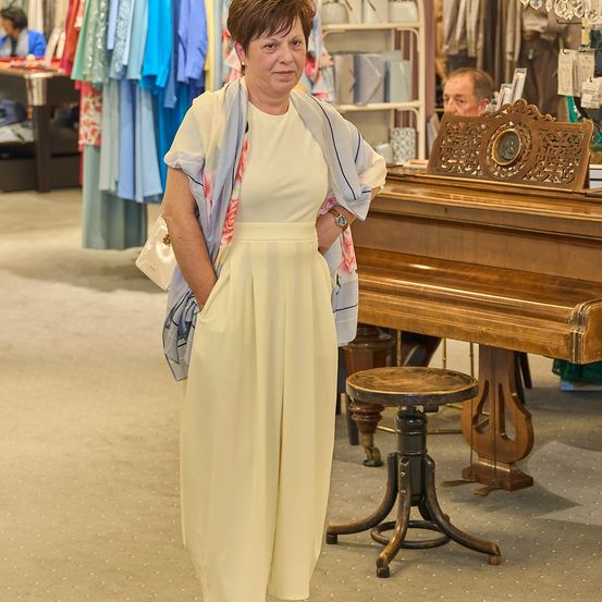 Eine Frau in einem gelben Kleid steht in einem Bekleidungsgeschäft, mit einem Klavier und einem Mann im Hintergrund.