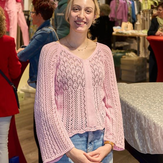 Eine Frau in einem rosa Spitzenoberteil und Jeans steht in einem Bekleidungsgeschäft und posiert für ein Foto. Hinter ihr sind eine Frau in einer Jeansjacke und eine Person in einem roten Oberteil zu sehen, zusammen mit einem Tisch mit verschiedenen Artikeln.