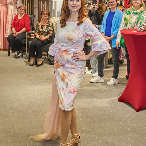 Eine Frau in einem Blumenkleid posiert in einem Bekleidungsgeschäft, mehrere Frauen im Hintergrund, einige sitzend und andere stehend.