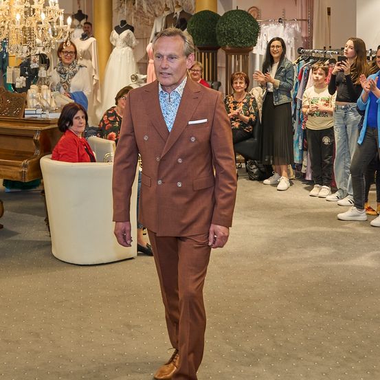 Ein Mann in einem braunen Anzug geht durch eine Boutique, wobei mehrere Menschen hinter ihm stehen und applaudieren.