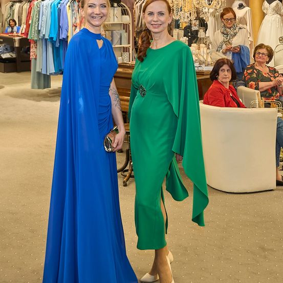 Zwei Frauen in eleganten Kleidern stehen in einem Boutique, eine in Blau und die andere in Grün, während zwei Frauen im Hintergrund sitzen, eine trägt ein rotes Shirt.