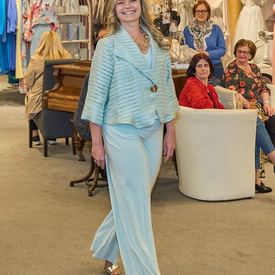 Eine Frau in einem hellblauen Anzug steht in einem Boutique, lächelt. Hinter ihr sitzt eine Frau in einem roten Mantel in einem weißen Stuhl. Andere Frauen sind sitzend und stehend umher.