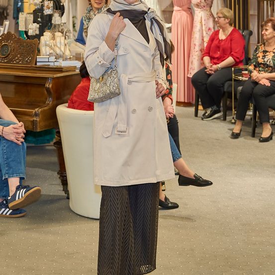 Eine Frau in einem Trenchcoat steht in einem Boutique, umgeben von sitzenden Frauen und einem hölzernen Klavier.