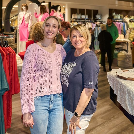 Zwei Frauen stehen in einem Bekleidungsgeschäft, eine in einem rosa Pullover und die andere in einem blauen Shirt, lächeln für ein Foto. Dahinter präsentieren Mannequins Kleidung und Leute stöbern.