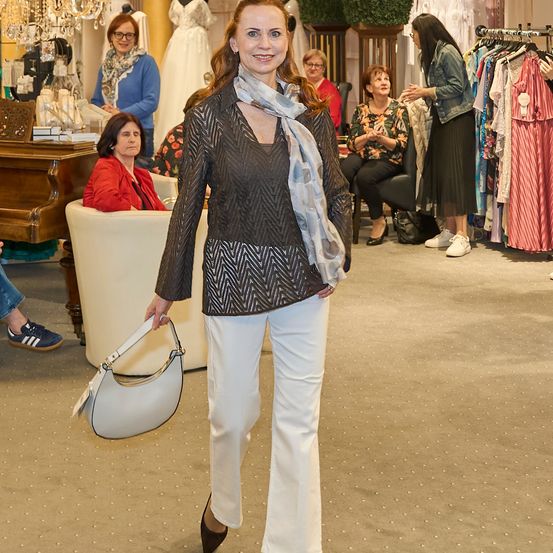 Eine Frau in einer schwarz-weiß gemusterten Bluse und weißen Hosen geht durch ein Bekleidungsgeschäft und hält eine weiße Handtasche. Hinter ihr sitzen und stehen mehrere Frauen, und Kleiderständer hängen an der Wand.