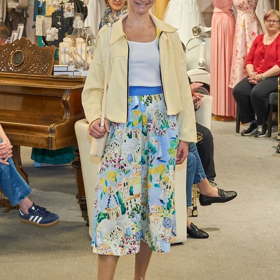 Eine Frau in einem Blumenrock und einer gelben Jacke steht in einem Bekleidungsgeschäft, lächelt und hält eine weiße Handtasche. Hinter ihr sind ein Klavier und eine Frau in einem roten Shirt zu sehen.