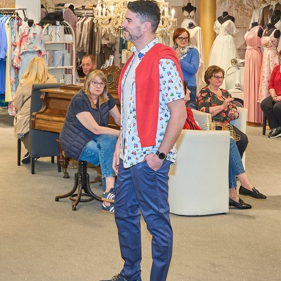 Ein Mann steht in einem Bekleidungsgeschäft, trägt eine rote Weste über einem gemusterten Hemd und blaue Jeans. Hinter ihm sitzen mehrere Frauen auf Stühlen und beobachten, während Mannequins im Hintergrund Kleider präsentieren.