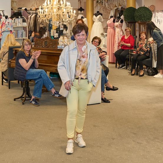 Eine Frau steht in einem Bekleidungsgeschäft, umgeben von Mannequins und Kunden. Sie trägt eine helle Jacke und hellgelbe Hosen.