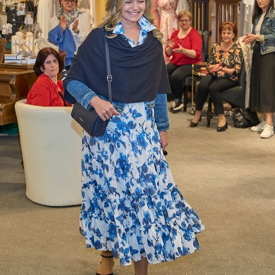 Eine Frau in einem blauen Blumenrock und einer Jeansjacke geht lächelnd in einem Geschäft, während andere um sie herum sitzen und stehen.