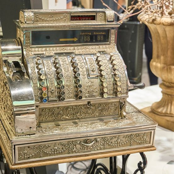 Eine verzierte alte Registrierkasse mit einem digitalen Display, das den Betrag von 18,08 anzeigt. Die Registrierkasse hat einen silbernen Körper und goldene Akzente.