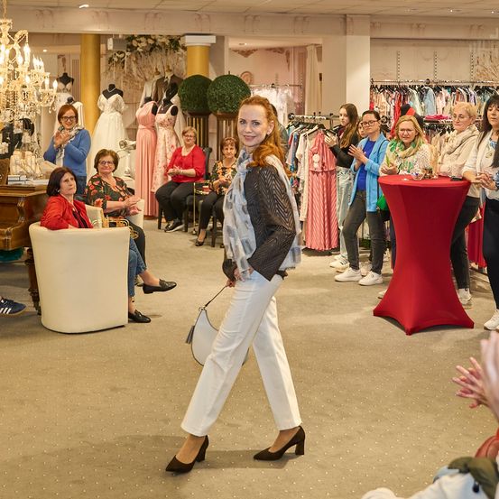 Eine Frau geht durch ein Modegeschäft, umgeben von sitzenden Frauen und einem roten Tisch mit Gegenständen. Im Hintergrund befinden sich Mannequins und Kleiderständer.