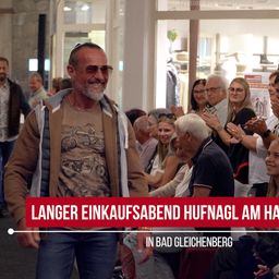 Ein Mann mit Bart und Sonnenbrille steht vor einer Gruppe von Menschen. Sie alle lächeln und klatschen. Der Mann trägt eine braune Jacke mit einem Motorrad-Aufdruck. Der Text auf dem Bild lautet 'Langer Einkaufsabend Hufnagel am Ha'.
