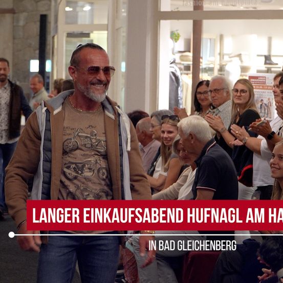 Ein Mann mit Bart und Sonnenbrille steht vor einer Gruppe von Menschen. Sie alle lächeln und klatschen. Der Mann trägt eine braune Jacke mit einem Motorrad-Aufdruck. Der Text auf dem Bild lautet 'Langer Einkaufsabend Hufnagel am Ha'.