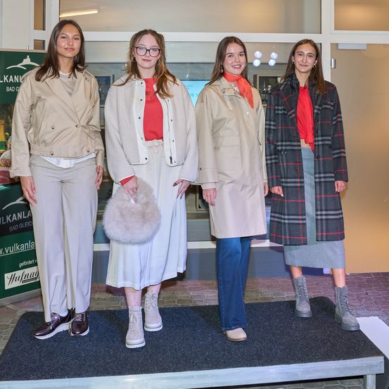 Vier Frauen stehen auf einer Bühne und tragen verschiedene Mäntel und Stiefel. Sie lächeln und scheinen Models zu sein. Die Frau auf der linken Seite hält eine Pelzhandtasche. Hinter ihnen ist eine Glaswand mit einem Schild.