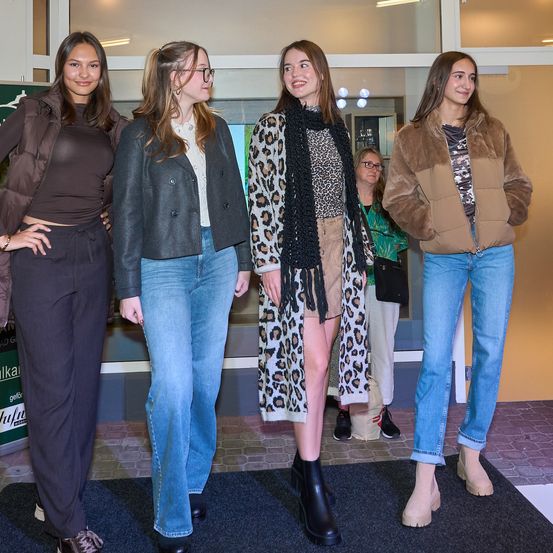 Fünf Frauen in modischen Outfits stehen auf einem schwarzen Teppich und posieren für ein Foto. Eine trägt einen Leopardenmantel, eine andere eine braune Daunenjacke. Hinter ihnen beobachtet eine Frau mit Brille.