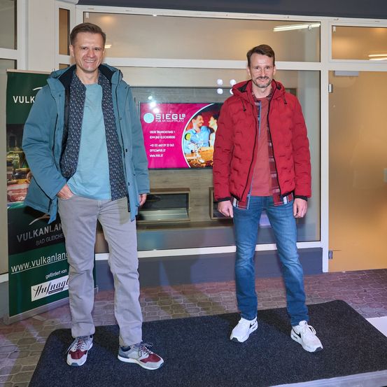 Zwei Männer stehen auf einem schwarzen Teppich vor einer Glastür. Der Mann auf der linken Seite trägt eine blaue Jacke und graue Hose, während der Mann auf der rechten Seite eine rote Jacke und blaue Jeans trägt. Hinter ihnen befindet sich ein Schild mit dem Text 'VULKAN' und einer Website-Adresse.