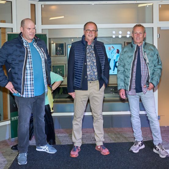 Drei Männer stehen vor einer Glastür und lächeln für ein Foto. Der Mann auf der linken Seite trägt ein blaues Hemd und eine Jacke, der Mann in der Mitte trägt eine blaue Weste über einem Hemd mit Blumenmuster und der Mann auf der rechten Seite trägt eine grüne Jacke. Hinter ihnen zeigt ein Fernsehbildschirm ein Bild von Menschen.