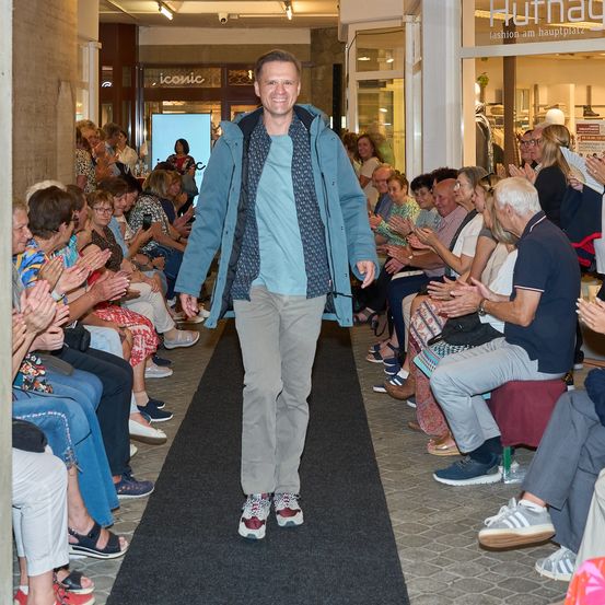 Ein Mann geht in einer blauen Jacke und khakifarbenen Hose über einen Laufsteg, während eine Menge Menschen hinter ihm applaudiert.