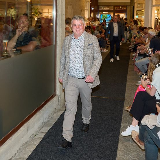 Ein Mann in einem Anzug geht einen Teppichgang in einem Einkaufszentrum entlang, mit Menschen, die dahinter sitzen und stehen.