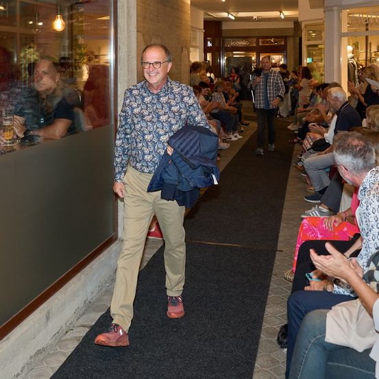 Ein Mann in einem gemusterten Hemd und khakifarbenen Hosen geht einen teppichbelegten Gang entlang und trägt eine blaue Jacke. Hinter ihm sitzen einige Leute in Stühlen, einige klatschen. Ein Mann mit einem Gehstock steht in der Nähe des Eingangs.
