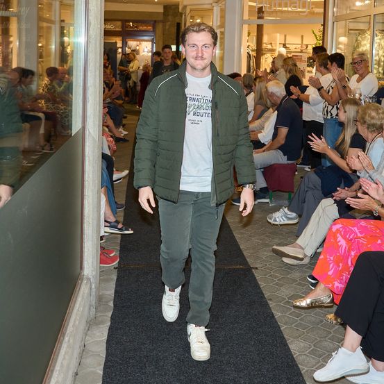 Ein Mann in einer grünen Jacke und weißen Sneakers geht durch einen Flur, umgeben von sitzenden Menschen, die applaudieren.