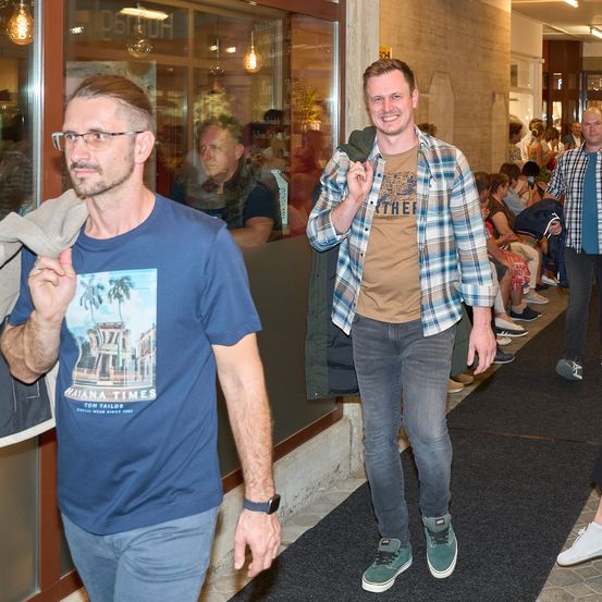 Zwei Männer gehen einen Flur hinunter, während im Hintergrund eine Gruppe sitzender Menschen zu sehen ist. Einer trägt ein blaues T-Shirt mit tropischem Druck, der andere ein kariertes Hemd.