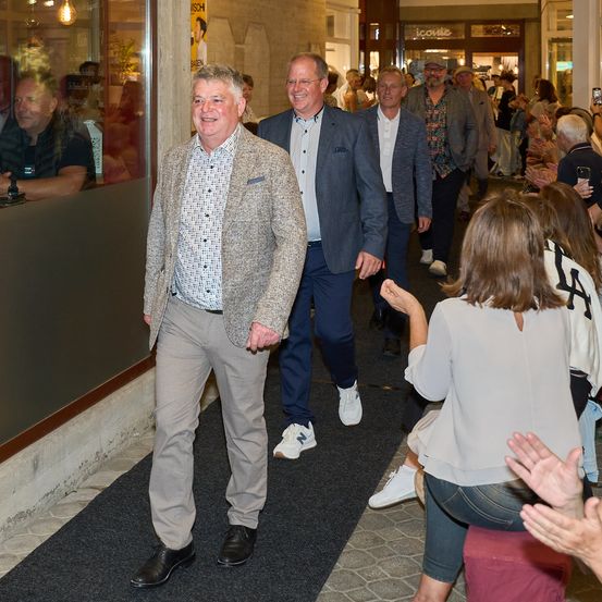Eine Gruppe von Menschen geht einen Flur entlang, wobei ein Mann vorne einen grauen Blazer trägt und lächelt. Dahinter gehen zwei andere Männer, einer trägt einen grauen Anzug. Rechts klatscht eine Frau.