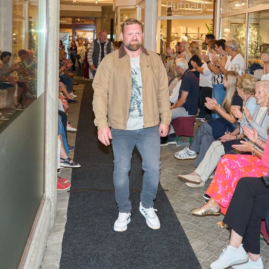 Ein Mann in einer braunen Jacke und Jeans geht einen Teppichgang hinunter, während ein Publikum auf beiden Seiten klatscht.