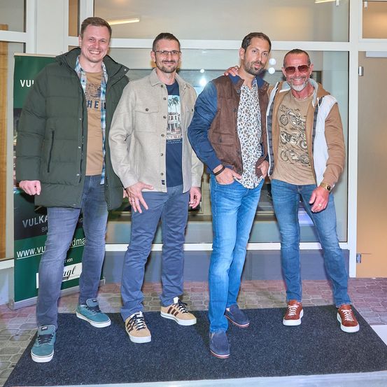 Vier Männer stehen auf einem schwarzen Teppich und lächeln für ein Foto. Sie tragen Freizeitkleidung und Turnschuhe. Hinter ihnen ist eine Glaswand mit einem Schild und einer Tür.