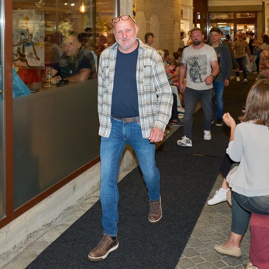 Ein Mann mit Brille und kariertem Jackett geht durch einen Flur, hinter ihm eine Menge Menschen.