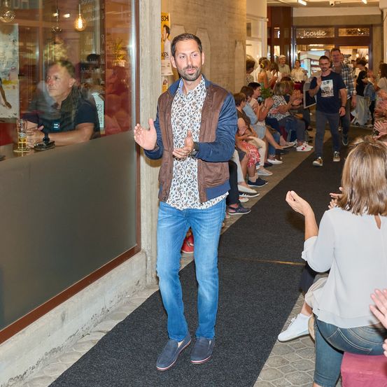 Ein Mann in einer braunen Jacke und blauen Jeans geht auf einem schwarzen Teppich in einem Flur, mit Menschen auf der Seite, die klatschen.