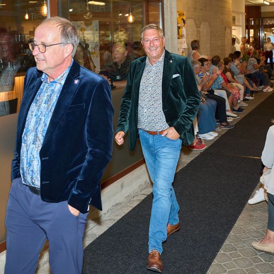 Zwei Männer gehen einen Teppichgang entlang, einer in einem blauen Samtjackett und der andere in einem grünen Samtjackett, mit einer Menge sitzender Menschen im Hintergrund.