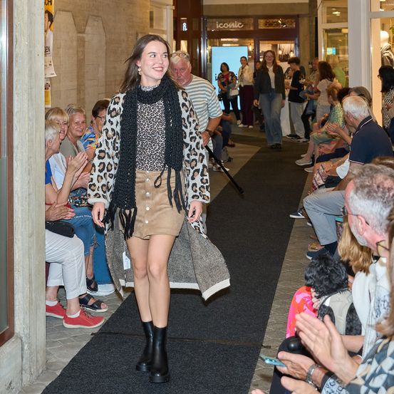 Eine Frau in einem Leopardenmustermantel und Rock geht eine Laufsteg hinunter, während ein Publikum zuschaut.