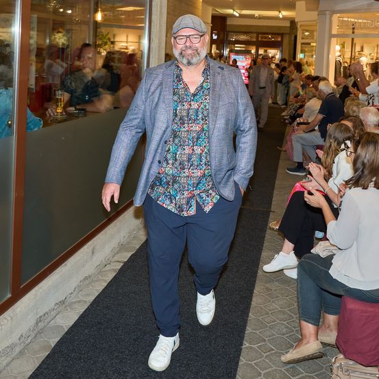 Ein Mann in einem Blumenhemd und Blazer geht einen Teppichgang hinunter, mit sitzenden und klatschenden Menschen im Hintergrund.