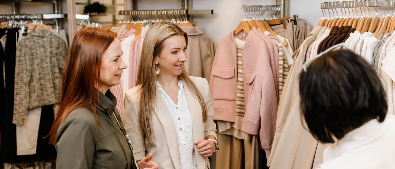 Zwei Frauen stehen vor einem Bekleidungsgeschäft, eine trägt einen beigefarbenen Blazer und die andere eine grüne Jacke, sie schauen sich Kleider an den Kleiderbügeln an.