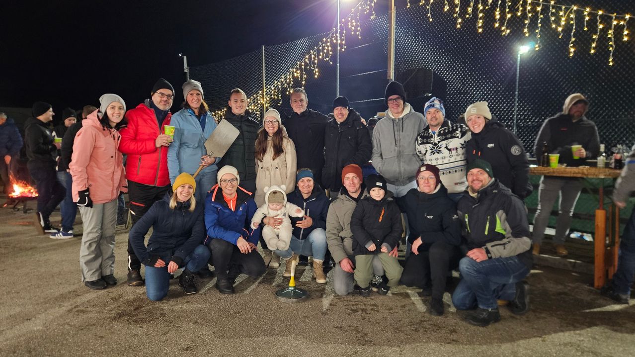 Eine Gruppe von Menschen in Winterkleidung, die ein Foto mit einem Baby in der Mitte, einem gelb-weißen Objekt und einem Zaun mit Lichtern macht.