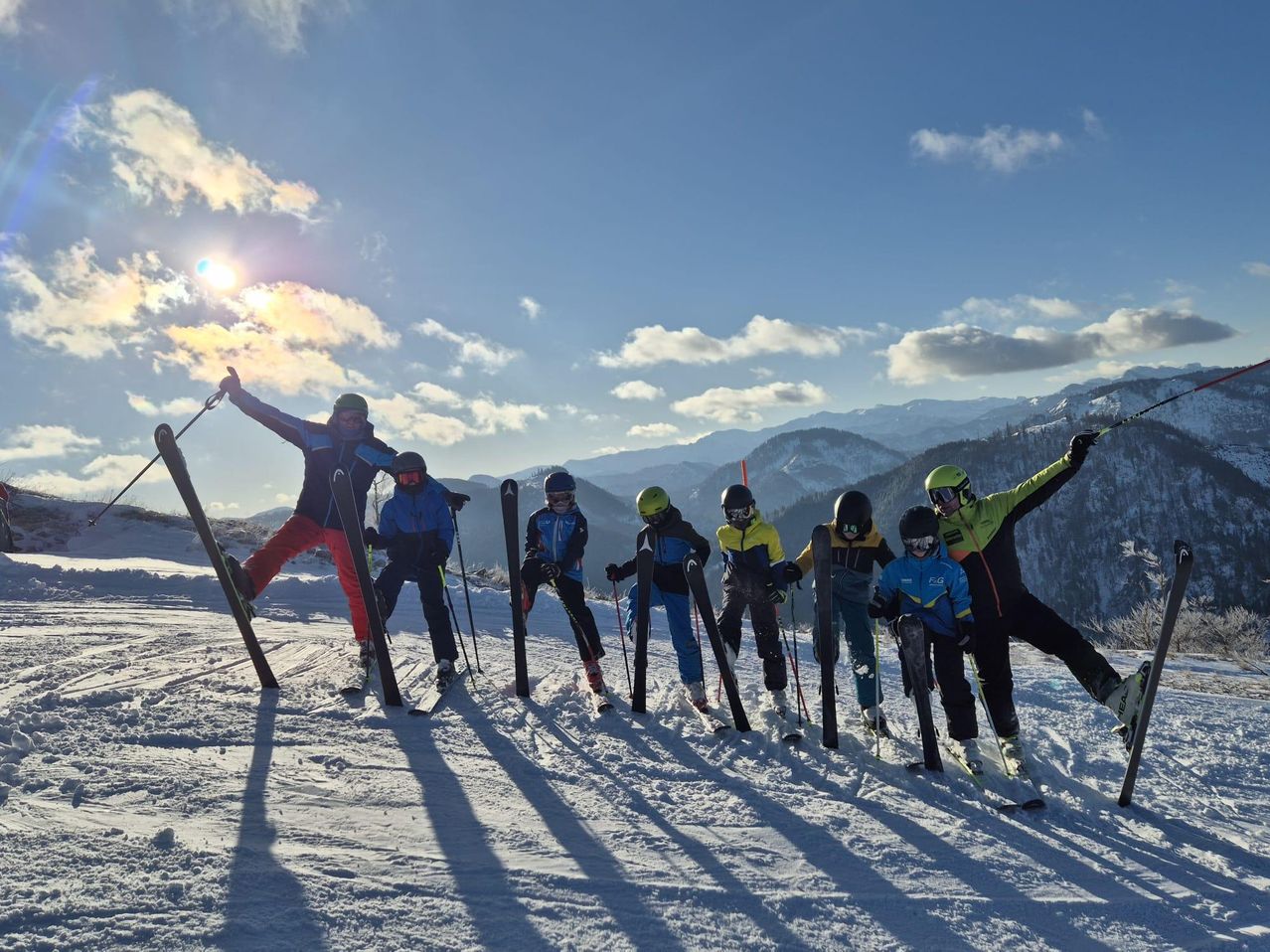 Eine Gruppe von Skifahrern, einschließlich Kindern, posiert für ein Foto auf einem verschneiten Berg. Sie tragen Winterkleidung und Skiausrüstung. Die Sonne scheint hell in der Ferne.