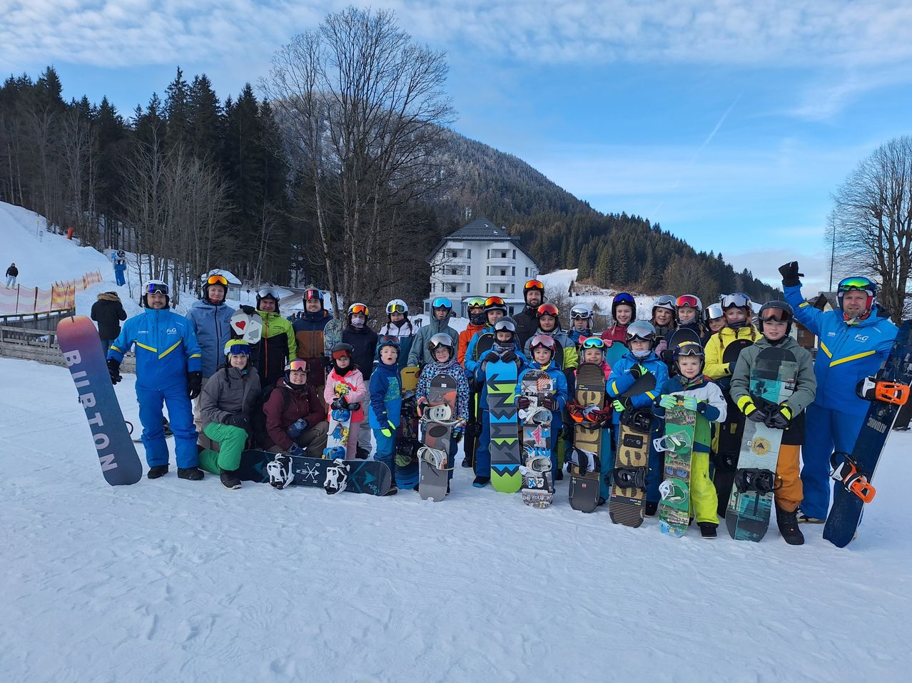 A group of snowboarders poses for a photo in the snow, with mountains and a building in the background.