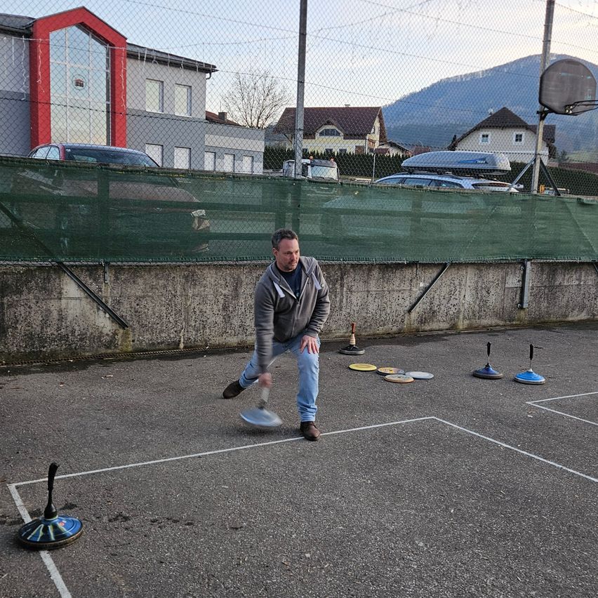 Ein Mann wirft einen Frisbee auf einem Spielfeld mit mehreren Hindernissen, in der Nähe eines Zauns mit geparkten Autos und Gebäuden.
