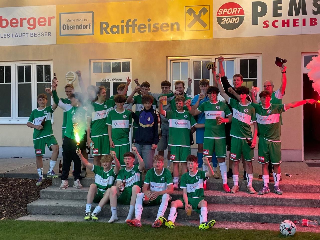 Eine Fußballmannschaft in grünen Uniformen posiert für ein Foto vor einem Gebäude mit einem Raiffeisen-Schild. Sie halten Getränke und einen grünen Leuchtkörper.